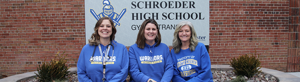Three women sitting on the edge of a large planter box with a sign behind them that reads Schroeder High School.