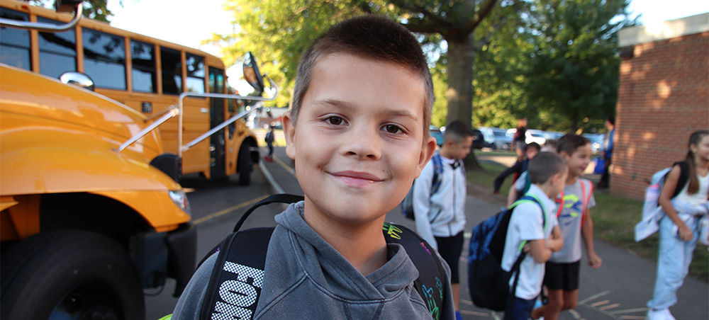 Boy wearing backpack with school buses and other kids wearing backpacks behind him.