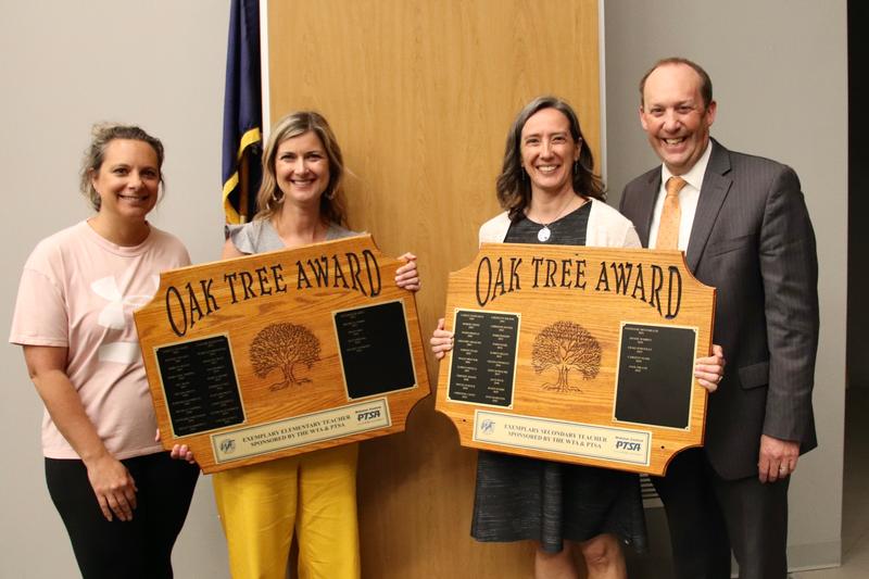Three women and a man standing together with the two women in the middle holding large plaques that read Oak Tree Award.