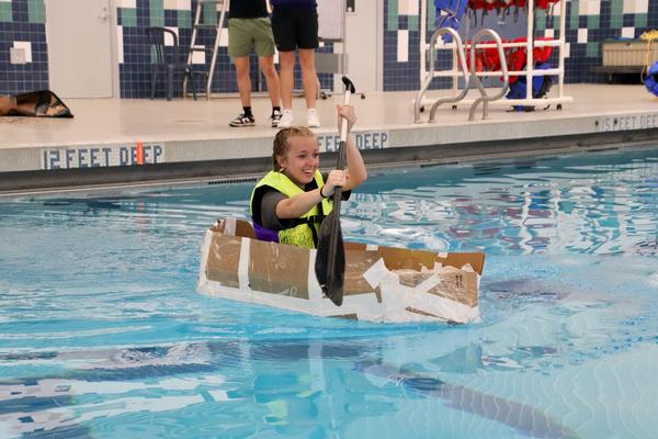 Teen paddling cardboard canoe in pool.
