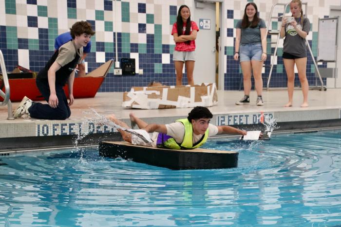 Teen laying on a flat cardboard boat and wearing cardboard hand fins to paddle in a pool.
