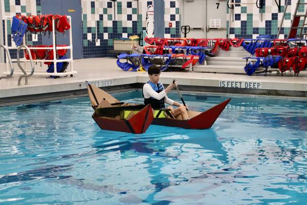 Teen paddling a cardboard outrigger canoe in a pool.
