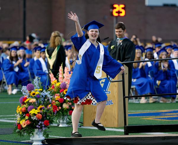 A high school student waving while wearing a royal blue graduation gown and cap with a white sash with the letter "S"