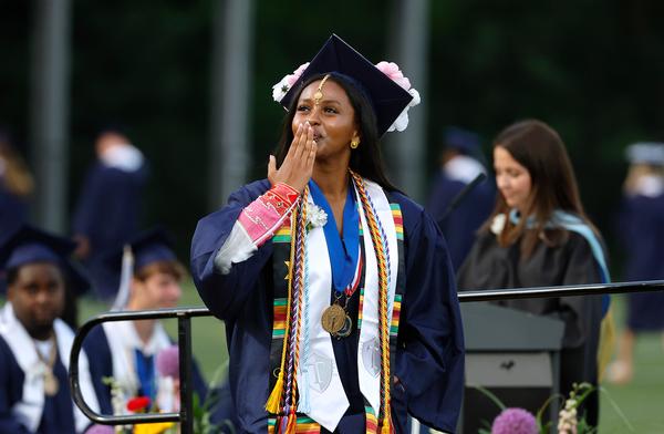 A high school girl blowing a kiss to someone while wearing a navy graduation gown and cap and a white sash with a silver T on it.