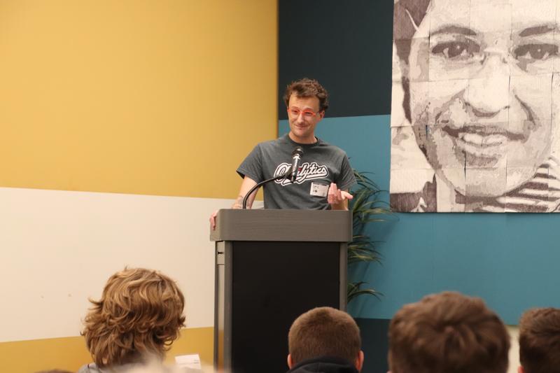 Man at podium talking. Backs of teen heads can be seen listening below him.