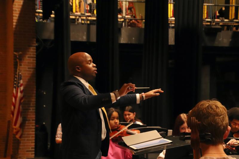 Man holding a baton conducting orchestra of teens.