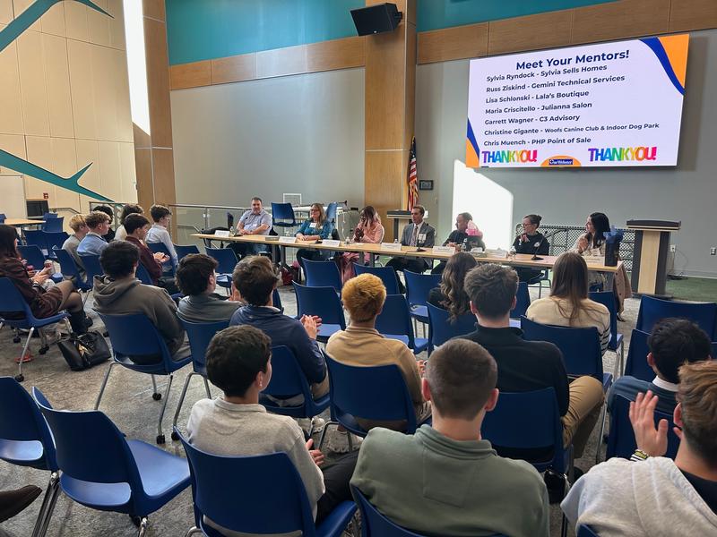 A large group of students seated toward a panel of seven adults. 