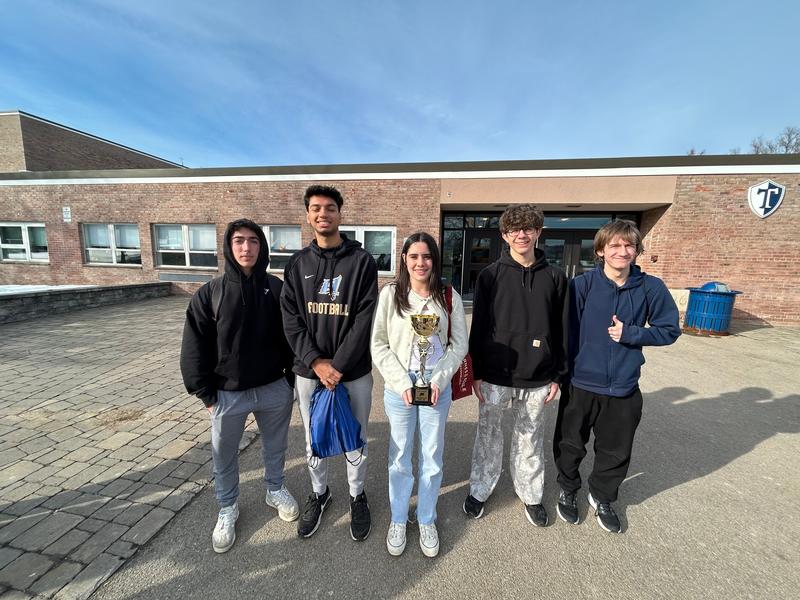 Four teen boys and a teen girl standing in front of a school.  The girl is holding a trophy.