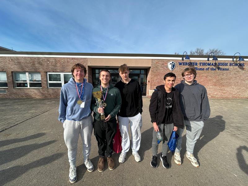 Three teen boys standing in front of a school. Two are wearing medals and one is hold a trophy.