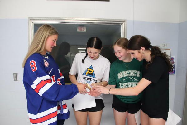 Young woman in red, white and blue jersey holding a gold medal out to three awe-struck teens.