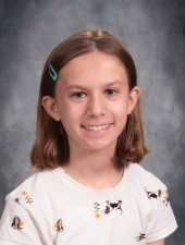 Smiling girl with teal barrette and wearing white shirt with yellow flowers scattered across it.