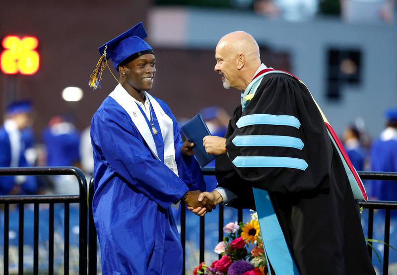 Teen in cap and gown receives diploma from adult.