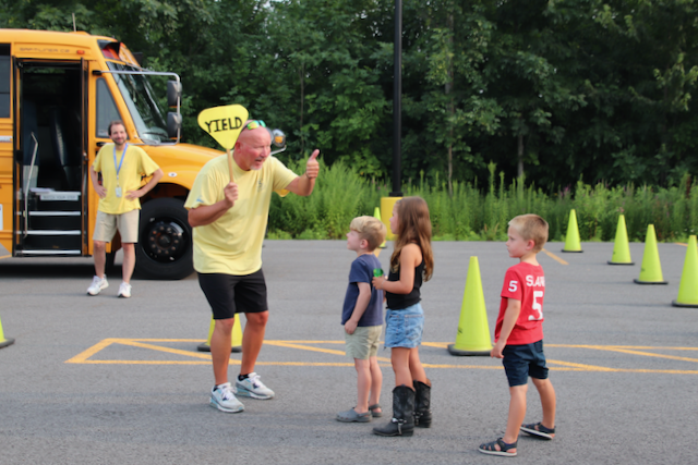 Man in yellow shirt holding Yield sign giving thumbs up to three small children. School bus in the background.