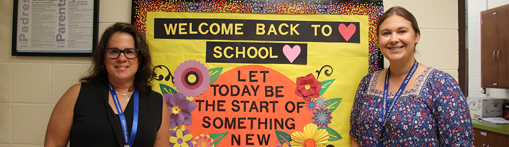 Two teachers standing byt bulletin board that says, "Welcome Back to School"