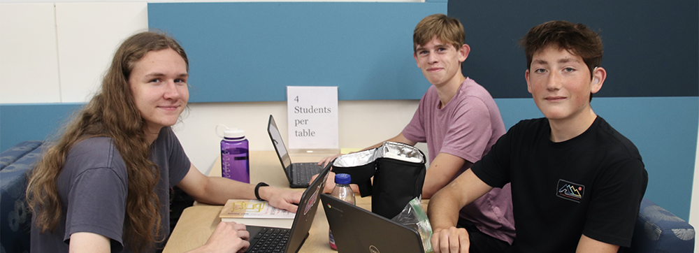 Three high school students sitting at a table with their laptops open.