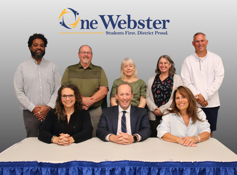 Four women and four men behind a table with the One Webster logo over them.