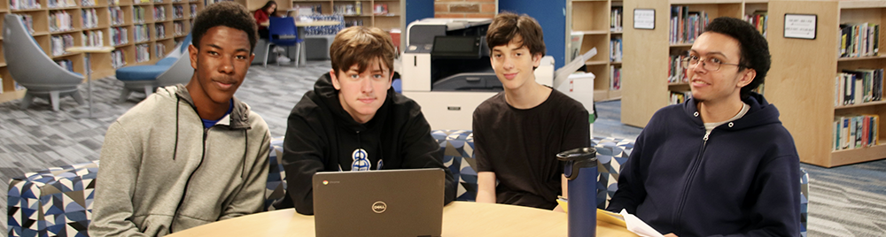 Four teens working over laptop at table.